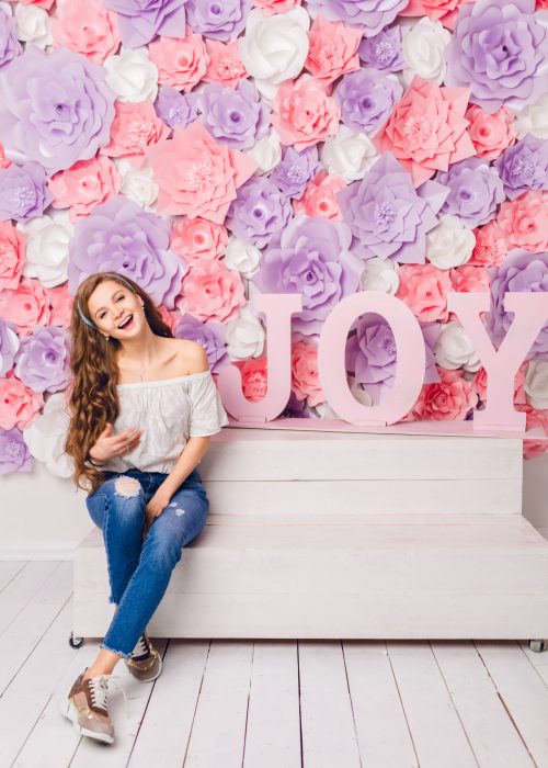 Cute blond girl sitting on a bench in a studio smiling widely. She has long curly hair and wears blue jeans and white t-shirt off the shoulder.She has pink background covered in flowers with word JOY.