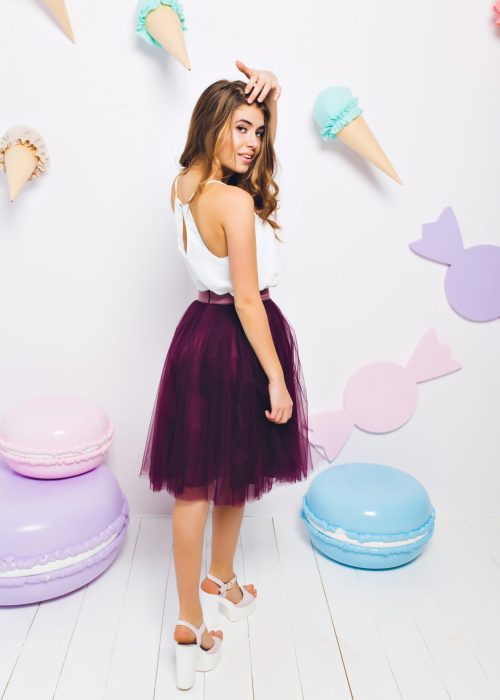 Slim tanned girl in long violet skirt looking over her shoulder posing on theme party and touching hair. Full-length portrait of gorgeous young woman standing in light room decorated with candies.